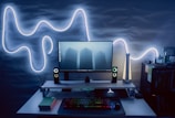 A modern computer setup with a flat-screen monitor displaying an image of three tall buildings in the fog. The setup includes LED-backlit speakers on either side of the monitor, and a backlit keyboard and mouse on the desk. On the left side, there is a book titled 'Do Epic Shit.' The wall behind the setup features an illuminated, abstract light design, adding an aesthetic glow to the scene. To the right, there's a shelf with various small objects and books, enhancing the room's ambience.