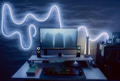A modern computer setup with a flat-screen monitor displaying an image of three tall buildings in the fog. The setup includes LED-backlit speakers on either side of the monitor, and a backlit keyboard and mouse on the desk. On the left side, there is a book titled 'Do Epic Shit.' The wall behind the setup features an illuminated, abstract light design, adding an aesthetic glow to the scene. To the right, there's a shelf with various small objects and books, enhancing the room's ambience.