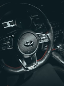 A close-up view of a car steering wheel, featuring the Kia logo at the center. The wheel is black with red stitching and has various control buttons on either side. The background is dark, highlighting the details of the steering wheel.