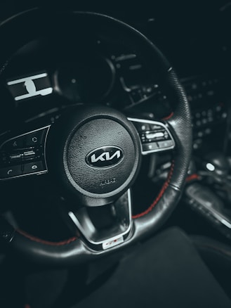 A close-up view of a car steering wheel, featuring the Kia logo at the center. The wheel is black with red stitching and has various control buttons on either side. The background is dark, highlighting the details of the steering wheel.