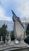 An intricately carved stone sculpture resembling a boat with a detailed mast and sail is positioned on a grave. Surrounding it are smaller stone ornaments including a decorative urn. In the background, there is a metal fence and trees with bare branches, under a partly cloudy sky.