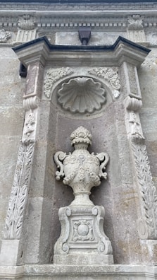 An ornate stone wall features a carved niche with intricate designs. Within the niche stands an elaborately decorated urn with floral motifs and scrollwork. Above the urn, a scallop shell relief is surrounded by decorative floral reliefs, adding to the detailed craftsmanship of the stone facade.