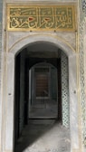 An architectural interior with intricate tile work on the walls and a decorative Arabic calligraphy panel above a stone archway. The corridor leads through a series of doorways, creating a sense of depth and perspective. The overall atmosphere appears historical and culturally rich.