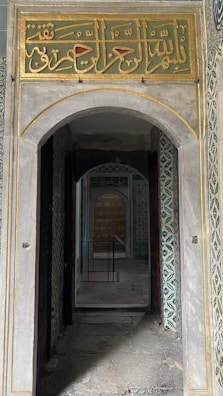 An architectural interior with intricate tile work on the walls and a decorative Arabic calligraphy panel above a stone archway. The corridor leads through a series of doorways, creating a sense of depth and perspective. The overall atmosphere appears historical and culturally rich.