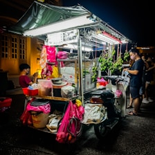 A street food cart is brightly illuminated at night. It is loaded with various items including hanging green mangoes and packages in colorful bags. Two people are nearby, one sitting and one standing, both wearing face masks. The cart is attached to a motorcycle, suggesting mobility.