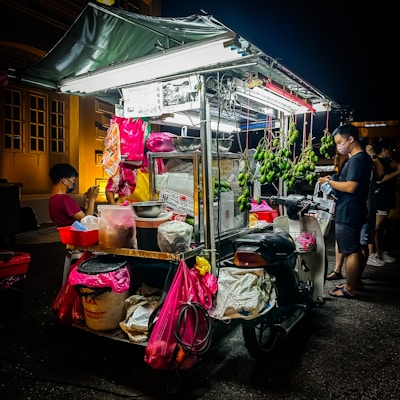 A street food cart is brightly illuminated at night. It is loaded with various items including hanging green mangoes and packages in colorful bags. Two people are nearby, one sitting and one standing, both wearing face masks. The cart is attached to a motorcycle, suggesting mobility.