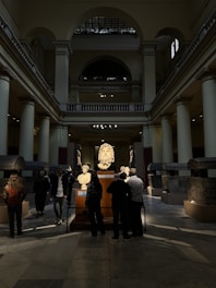 A spacious interior of a museum gallery with high ceilings and columns features several people observing sculptures and artifacts displayed on stands. The room is illuminated by a combination of natural light from windows and artificial spotlights highlighting the exhibits, while shadows and darker areas are present on the floor.