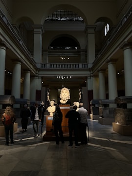 A spacious interior of a museum gallery with high ceilings and columns features several people observing sculptures and artifacts displayed on stands. The room is illuminated by a combination of natural light from windows and artificial spotlights highlighting the exhibits, while shadows and darker areas are present on the floor.