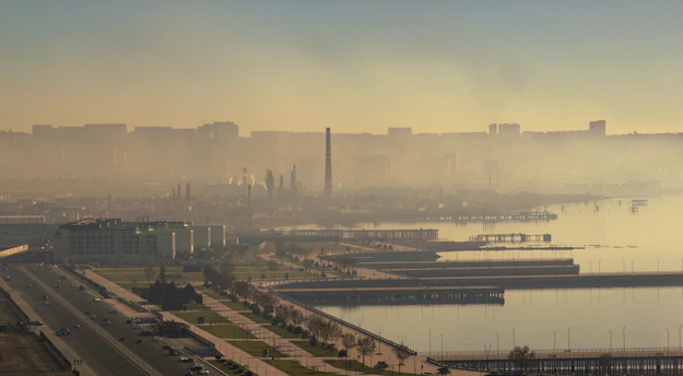 An industrial cityscape with a dense haze of smog enveloping the skyline, large chimneys emitting smoke in the background, and a waterfront with piers in the foreground. Multiple buildings line a major roadway that runs parallel to the water, while the muted colors of the sky blend with the urban environment.