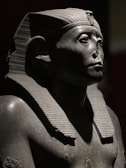 A stone sculpture of an ancient Egyptian pharaoh with a headdress, partially illuminated by dramatic lighting. The figure has a serene expression and is missing a portion of the nose.