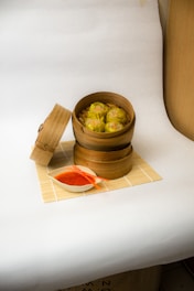 A bamboo steamer contains six pieces of siu mai dumplings, placed on a bamboo mat. The lid of the steamer is open and lies on its side next to it. Next to the steamer is a small white dish filled with red-orange sauce and a pair of orange chopsticks resting on the sauce bowl. The background is plain and light-colored, creating a clean and minimalistic setup.