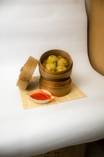 A bamboo steamer contains six pieces of siu mai dumplings, placed on a bamboo mat. The lid of the steamer is open and lies on its side next to it. Next to the steamer is a small white dish filled with red-orange sauce and a pair of orange chopsticks resting on the sauce bowl. The background is plain and light-colored, creating a clean and minimalistic setup.