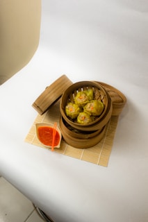 A bamboo steamer basket contains several pieces of yellow dumplings with orange toppings, placed on a bamboo mat. Beside it, there is a small dish of red sauce with a small orange utensil.