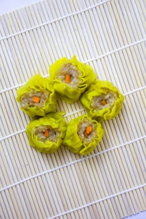 Five yellow dumplings arranged on a light bamboo mat. Each dumpling is filled with a meat-like filling and topped with a small piece of orange carrot.