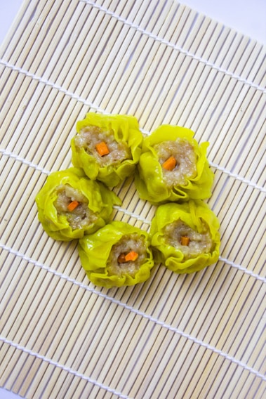 Five pieces of yellow dumplings with a filling that appears to contain minced meat and small orange carrot cubes, arranged neatly on a bamboo sushi mat. The dumplings are placed in a circular pattern, creating an aesthetically pleasing presentation.
