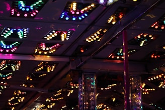 The image shows an interior ceiling adorned with variously colored, illuminated light fixtures. The light fixtures are arranged in patterns with several triangular or V-shaped designs. The lights are predominantly multicolored, creating a vibrant and lively atmosphere with reflections adding depth to the scene.