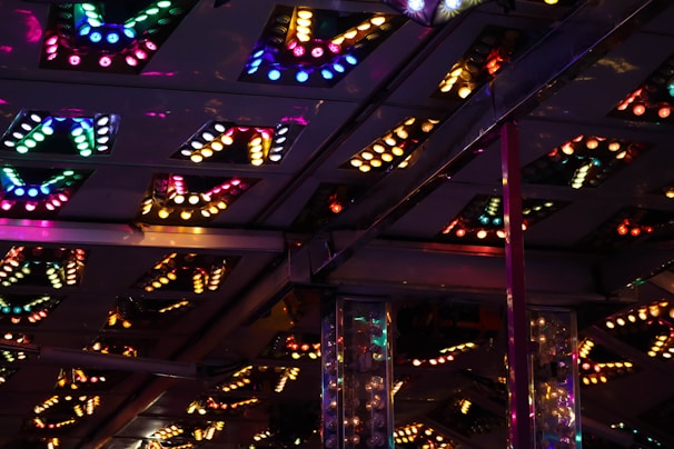The image shows an interior ceiling adorned with variously colored, illuminated light fixtures. The light fixtures are arranged in patterns with several triangular or V-shaped designs. The lights are predominantly multicolored, creating a vibrant and lively atmosphere with reflections adding depth to the scene.