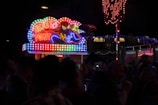 A vibrant, neon-lit booth in a dark setting prominently displays the words 'Ticket Point' with a dynamic superhero illustration. The colorful neon lights create a lively and energetic atmosphere, illuminating the immediate surroundings. In the foreground, silhouettes of people are visible, suggesting a bustling environment, possibly a fair or an event.