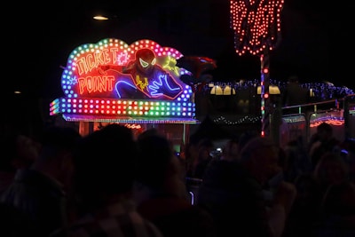 A vibrant, neon-lit booth in a dark setting prominently displays the words 'Ticket Point' with a dynamic superhero illustration. The colorful neon lights create a lively and energetic atmosphere, illuminating the immediate surroundings. In the foreground, silhouettes of people are visible, suggesting a bustling environment, possibly a fair or an event.