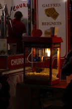 A popcorn machine illuminated from within, filled with freshly popped corn, sits in front of a colorful concession stand. To the left, a man in a red shirt appears to be preparing food. Brightly colored signs advertise 'Churros' and 'Gaufres Beignets,' with images of waffles displayed. The setting evokes the feel of a bustling fair or carnival.