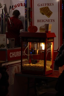 A popcorn machine illuminated from within, filled with freshly popped corn, sits in front of a colorful concession stand. To the left, a man in a red shirt appears to be preparing food. Brightly colored signs advertise 'Churros' and 'Gaufres Beignets,' with images of waffles displayed. The setting evokes the feel of a bustling fair or carnival.