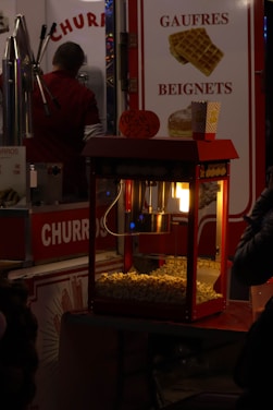 A popcorn machine illuminated from within, filled with freshly popped corn, sits in front of a colorful concession stand. To the left, a man in a red shirt appears to be preparing food. Brightly colored signs advertise 'Churros' and 'Gaufres Beignets,' with images of waffles displayed. The setting evokes the feel of a bustling fair or carnival.