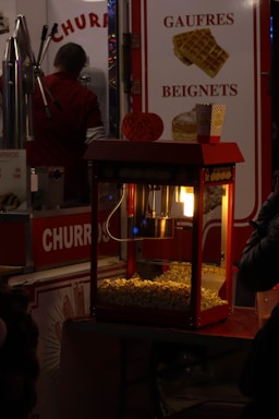 A popcorn machine illuminated from within, filled with freshly popped corn, sits in front of a colorful concession stand. To the left, a man in a red shirt appears to be preparing food. Brightly colored signs advertise 'Churros' and 'Gaufres Beignets,' with images of waffles displayed. The setting evokes the feel of a bustling fair or carnival.