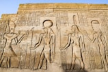 Ancient Egyptian hieroglyphics and carvings depicting figures of deities and historical figures. Intricate symbols and artwork are etched into the stone, showcasing traditional Egyptian attire and accessories.