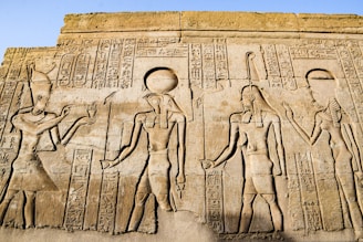 Ancient Egyptian hieroglyphics and carvings depicting figures of deities and historical figures. Intricate symbols and artwork are etched into the stone, showcasing traditional Egyptian attire and accessories.