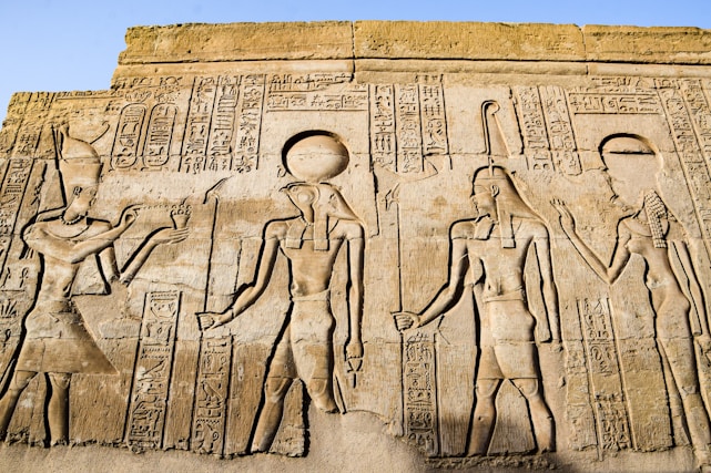 Ancient Egyptian hieroglyphics and carvings depicting figures of deities and historical figures. Intricate symbols and artwork are etched into the stone, showcasing traditional Egyptian attire and accessories.