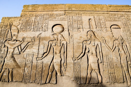 Ancient Egyptian hieroglyphics and carvings depicting figures of deities and historical figures. Intricate symbols and artwork are etched into the stone, showcasing traditional Egyptian attire and accessories.