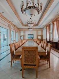 A spacious, elegantly decorated dining room with a long wooden table surrounded by intricately designed wooden chairs. The room features ornate chandeliers hanging from the ceiling, large windows with curtains, and polished marble flooring. The symmetrical arrangement creates a formal and grand atmosphere.