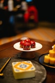 A close-up view of a dessert table featuring a white cake stand with four red glazed pastries. Next to it, there are several round and yellow cylindrical desserts on a black plate. In the foreground, a decorative object with intricate blue and orange patterns sits on a table. The background is blurred, indicating a warm, dimly-lit environment.