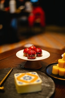 A close-up view of a dessert table featuring a white cake stand with four red glazed pastries. Next to it, there are several round and yellow cylindrical desserts on a black plate. In the foreground, a decorative object with intricate blue and orange patterns sits on a table. The background is blurred, indicating a warm, dimly-lit environment.