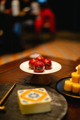 A close-up view of a dessert table featuring a white cake stand with four red glazed pastries. Next to it, there are several round and yellow cylindrical desserts on a black plate. In the foreground, a decorative object with intricate blue and orange patterns sits on a table. The background is blurred, indicating a warm, dimly-lit environment.