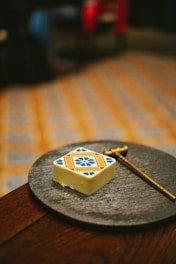 A decorative square piece of chocolate with a colorful geometric pattern featuring blue, orange, and red elements rests on a dark, round slate plate. A metallic tool resembling a small hammer or key lies next to the chocolate, placed on a rich wooden table. The background shows a blurred patterned floor with warm colors.