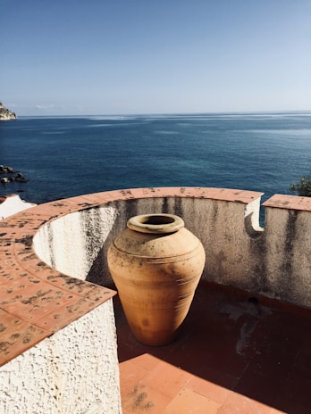 A large terracotta pot stands on a brick terrace overlooking a vast, calm blue sea. The terrace has a rough white curved wall with bricks lining the top, providing a warm contrast to the clear sky and water. Part of a rocky coastline is visible in the distance.