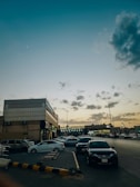 A street scene featuring colorful 2000s cars parked under neon signs at dusk.