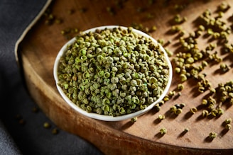 A small white bowl filled with green peppercorns is placed on a wooden surface. Some peppercorns are scattered around the bowl, adding to the rustic and organic feel of the scene. The lighting highlights the vibrant green color and texture of the peppercorns.