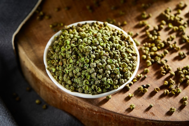 A small white bowl filled with green peppercorns is placed on a wooden surface. Some peppercorns are scattered around the bowl, adding to the rustic and organic feel of the scene. The lighting highlights the vibrant green color and texture of the peppercorns.