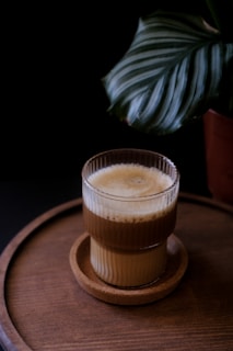 A layered coffee drink served in a clear, ribbed glass on a round wooden tray. A large green leaf partially frames the upper right corner, adding a touch of natural decor.