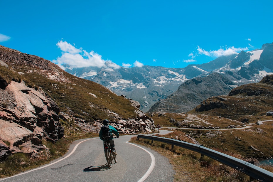 A cyclist is riding along a winding mountain road surrounded by rocky terrain and grassy slopes. Snow-capped mountain peaks tower in the background under a clear blue sky scattered with a few clouds.