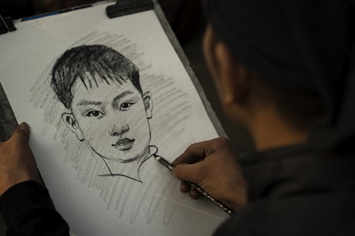 A person is sketching a portrait of a young boy with short hair on a sheet of paper using a pencil. The hand holding the pencil is seen in the foreground, and the artwork features shading and detailed lines.