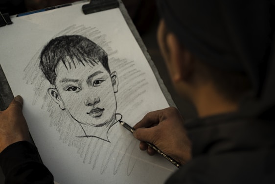 A person is sketching a portrait of a young boy with short hair on a sheet of paper using a pencil. The hand holding the pencil is seen in the foreground, and the artwork features shading and detailed lines.