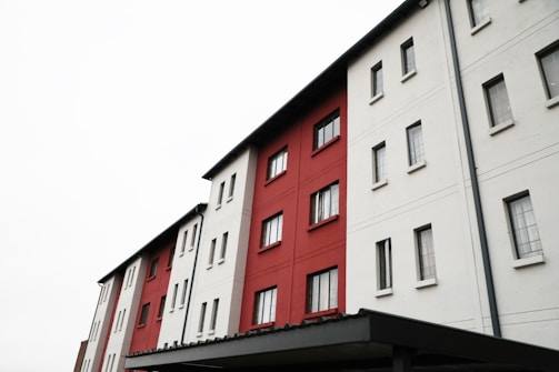 A multi-story building with a modern design features alternating sections of red and white walls. The building has multiple rectangular windows arranged in a grid-like pattern. The foreground includes part of a flat-roofed structure, potentially an overhang or entryway.