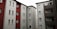 Several apartment buildings are arranged closely together. The structures have multiple floors with uniform windows, featuring varied colors including white, red, and grey on their facades. The scene is devoid of people, capturing only the exterior architectural design of the buildings.