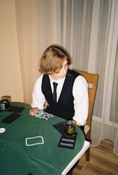 A person with shoulder-length hair is sitting at a poker table. They are wearing a white shirt, black vest, and tie, with an array of poker chips in front of them. There are two glasses with drinks on the table, and a smartphone is placed nearby. The background consists of vertical striped curtains.