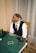 A person with shoulder-length hair is sitting at a poker table. They are wearing a white shirt, black vest, and tie, with an array of poker chips in front of them. There are two glasses with drinks on the table, and a smartphone is placed nearby. The background consists of vertical striped curtains.