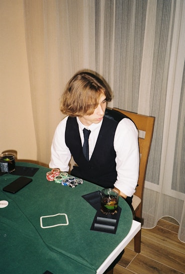 A person with shoulder-length hair is sitting at a poker table. They are wearing a white shirt, black vest, and tie, with an array of poker chips in front of them. There are two glasses with drinks on the table, and a smartphone is placed nearby. The background consists of vertical striped curtains.