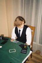 A person with shoulder-length hair is sitting at a poker table. They are wearing a white shirt, black vest, and tie, with an array of poker chips in front of them. There are two glasses with drinks on the table, and a smartphone is placed nearby. The background consists of vertical striped curtains.